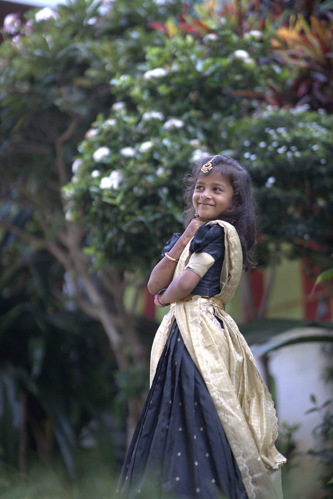 Chaithrika in a black and gold half-saree in a flowering garden, smiling shyly
