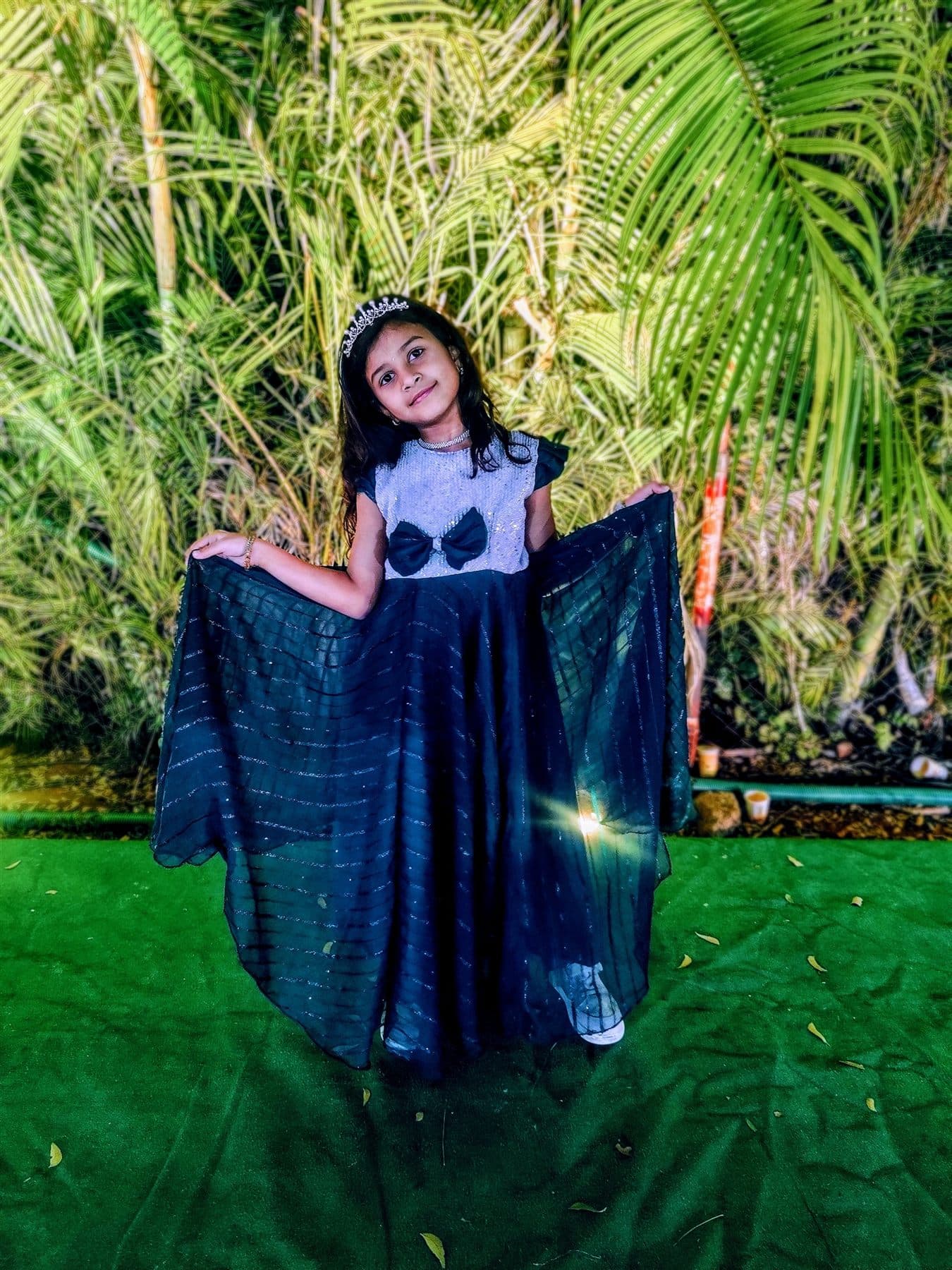 Chaithrika holding out a dark green gown, wearing a tiara, with palm leaves behind her