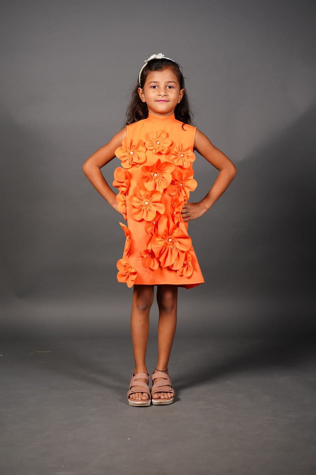 Chaithrika standing with hands on hips in an orange dress with three-dimensional flowers, against a grey studio backdrop