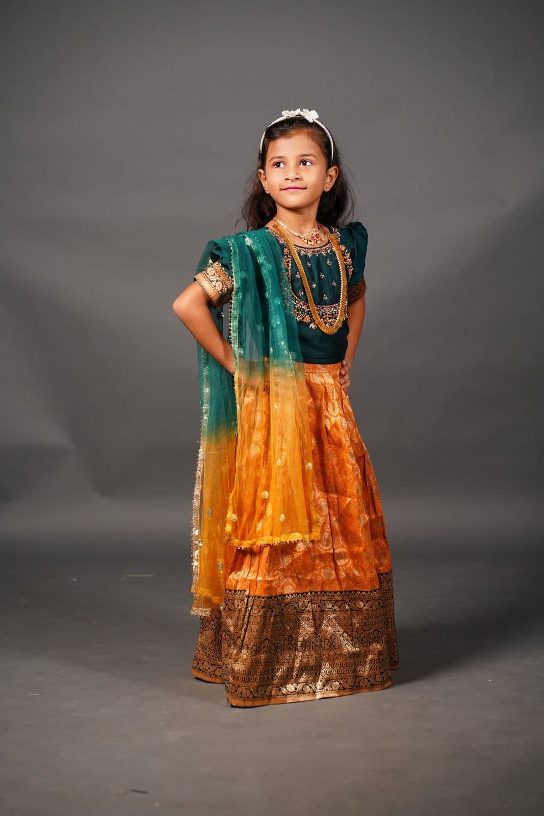 Chaithrika in a green blouse, orange silk lehenga and teal and yellow dupatta, looking off to one side