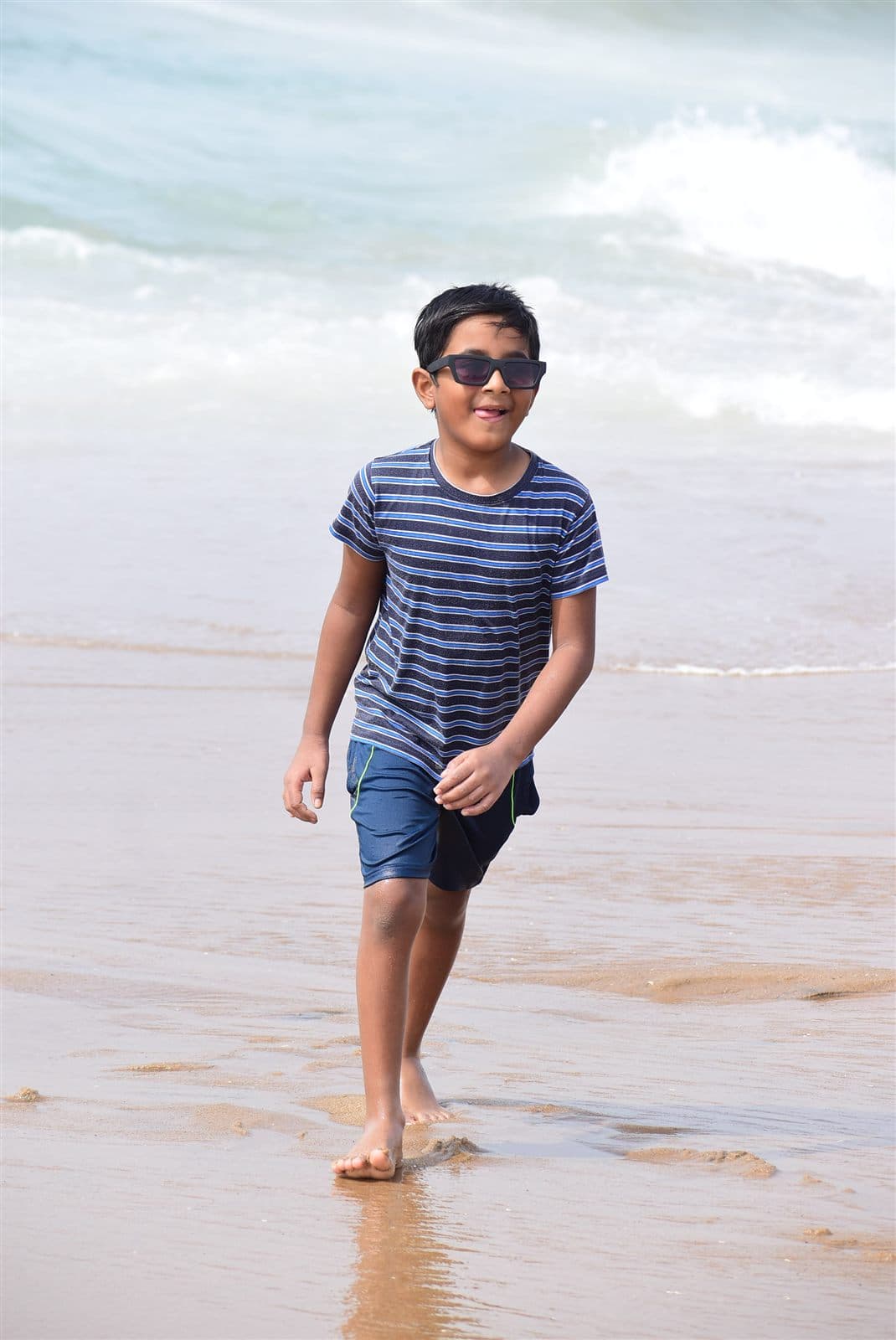 Nihan walking on the beach in a striped tee and sunglasses with waves behind