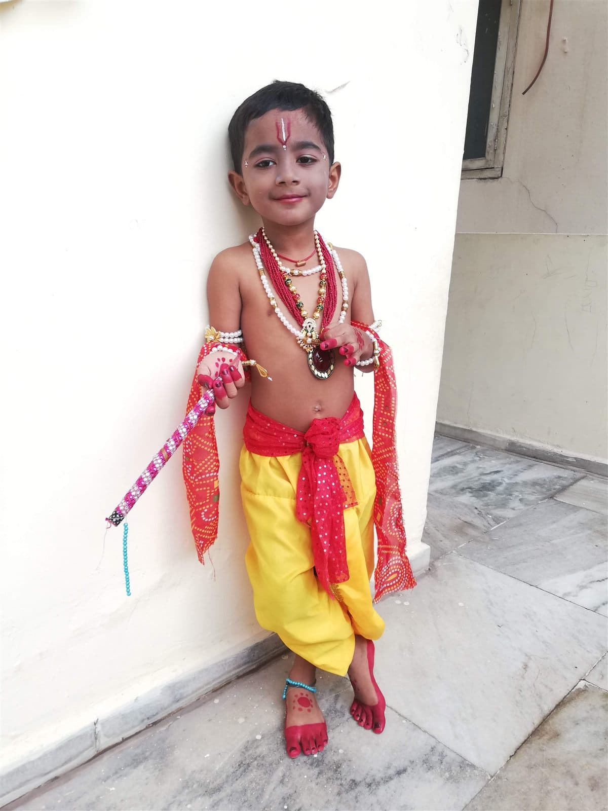 Young Nihan dressed as little Krishna in a yellow dhoti and red scarf against a white wall
