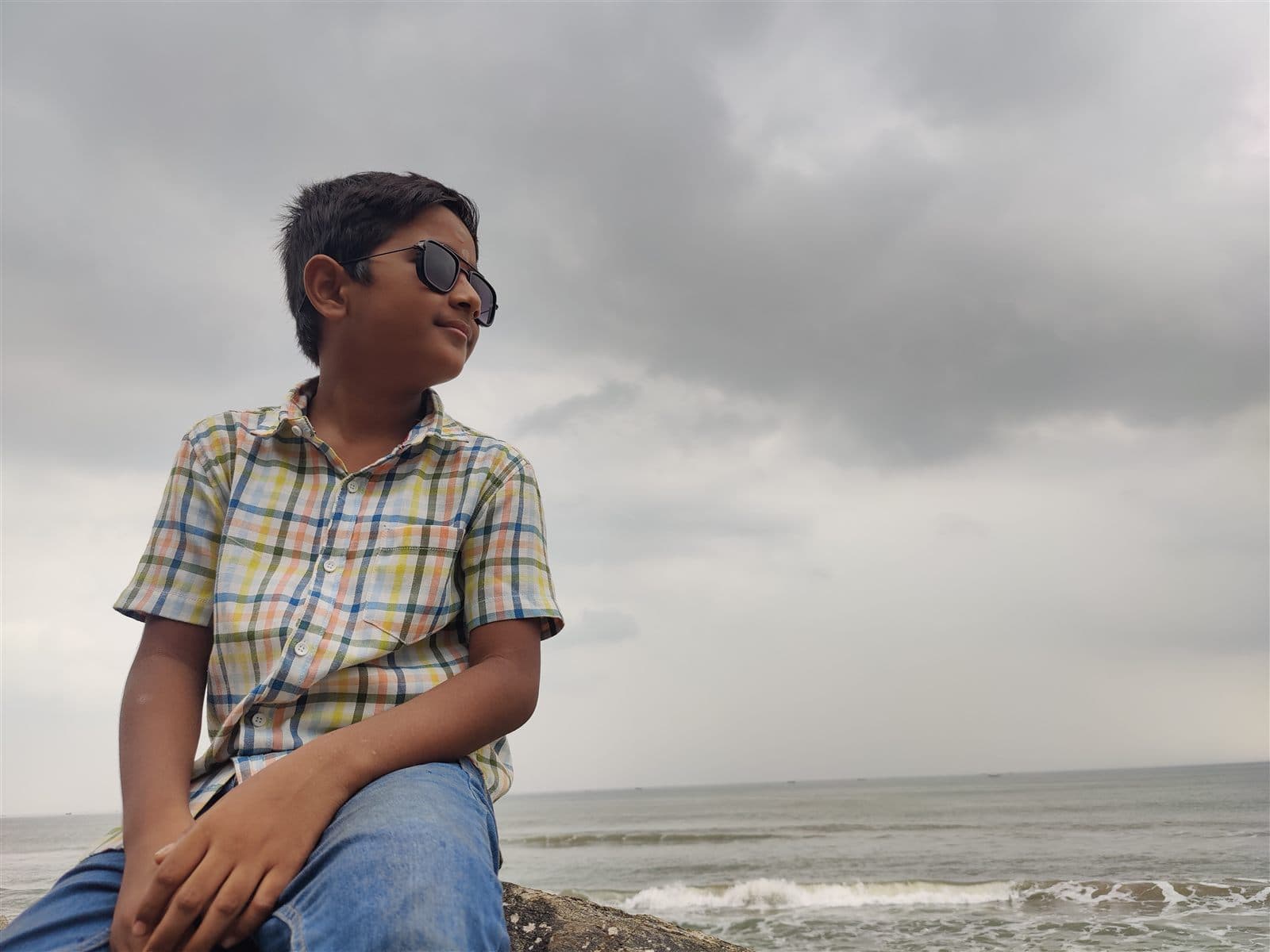 Nihan in a checked shirt and sunglasses, sitting on the rocks by the sea under a cloudy sky